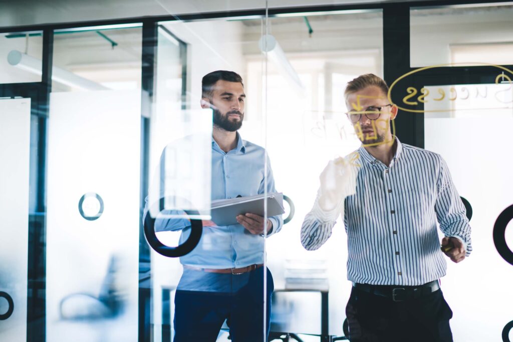 Two men working together at a clear glass board.