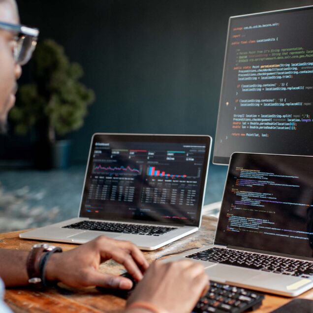 A man sitting at his desk working on two laptops and a large monitor.